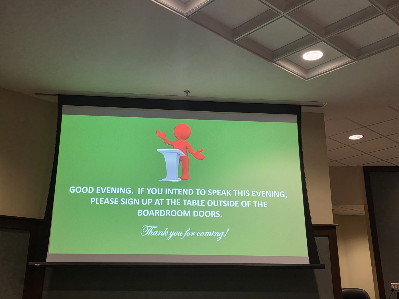 Boardroom screen reading: Good evening. If you intend to speak this evening, please sign up at the table outside of the boardroom doors.