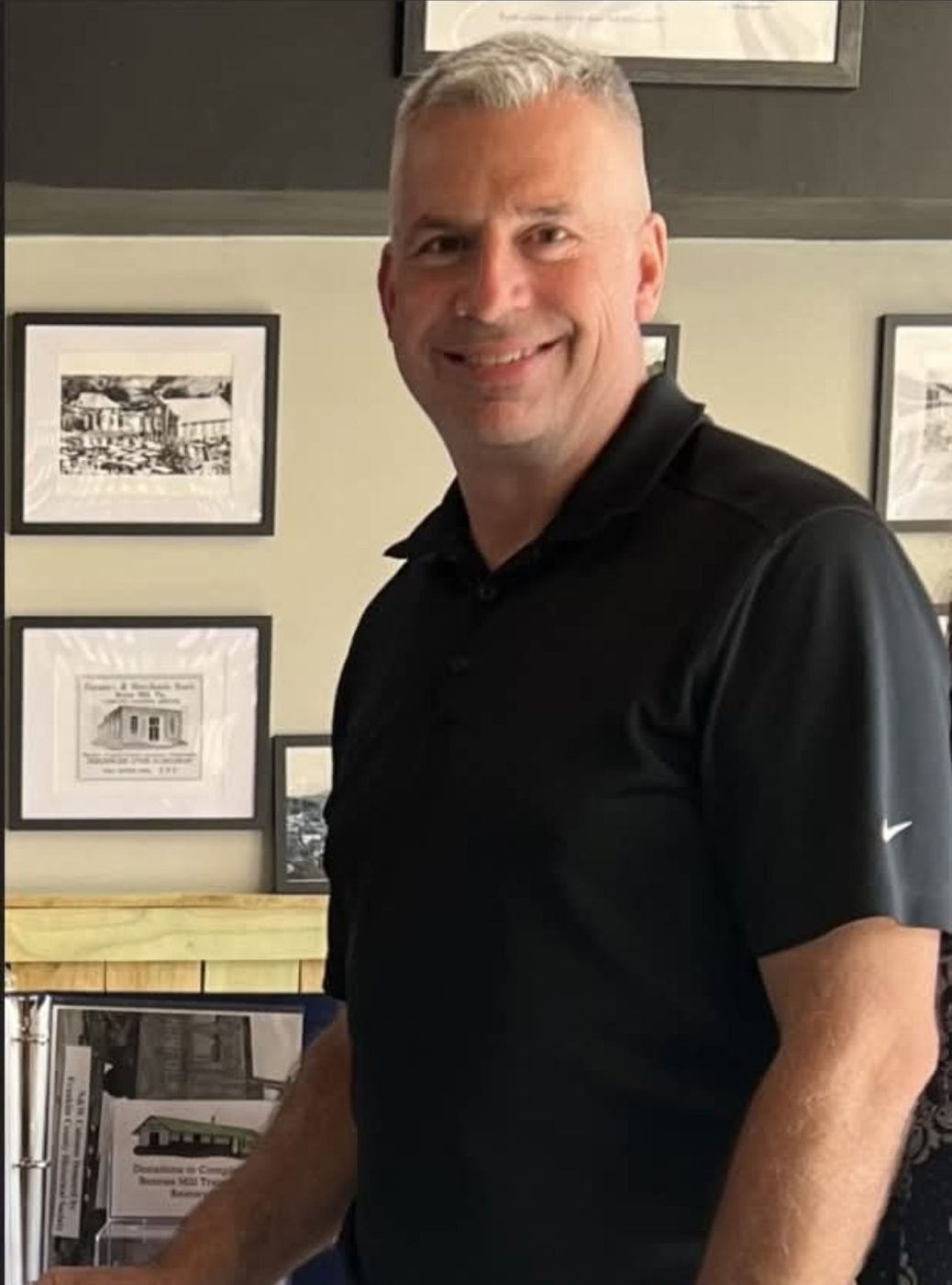 Mike Meredith smiling in a black polo shirt, standing in front of framed historical photographs